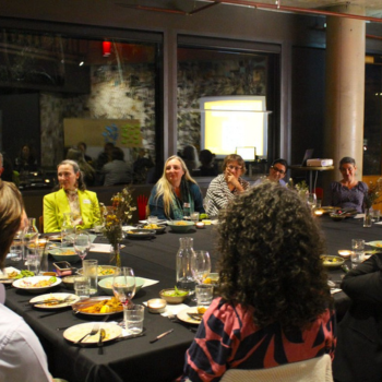 Group of people seated around a large dinner table at an indoor event, engaged in conversation with food and drinks.